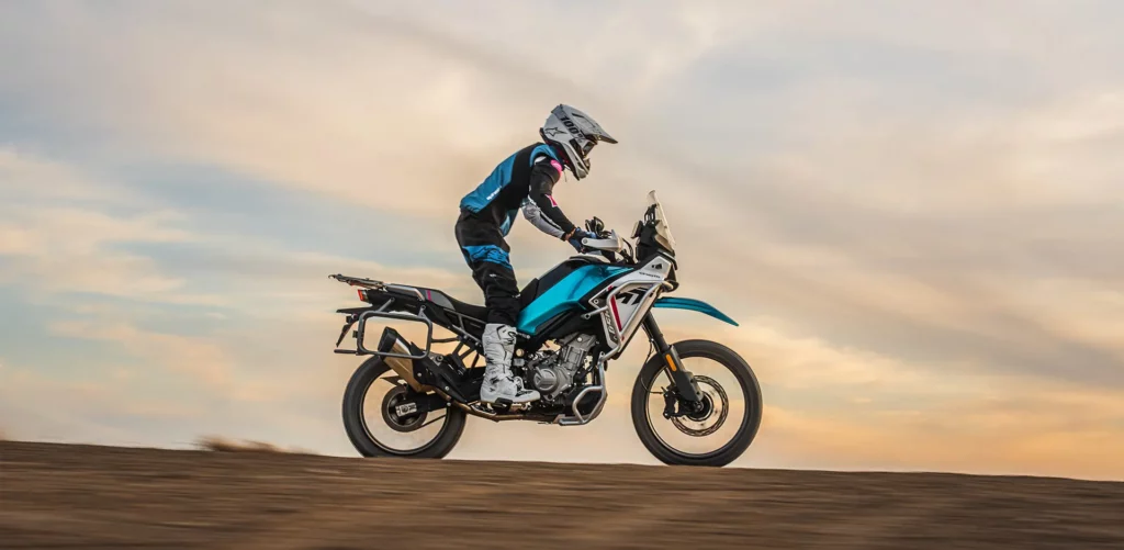 Ein Motorradfahrer in sportlicher Ausrüstung fährt auf einem CFMOTO Motorrad über eine sandige Landschaft bei Sonnenuntergang.